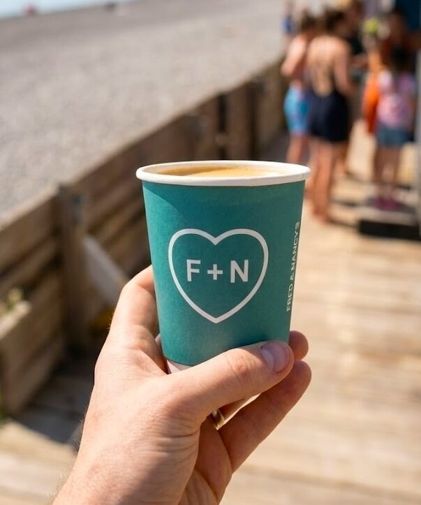 A Fred + Nancy's branded cup held in hand on Killiney Beach, with the queue at the Airstream behind.