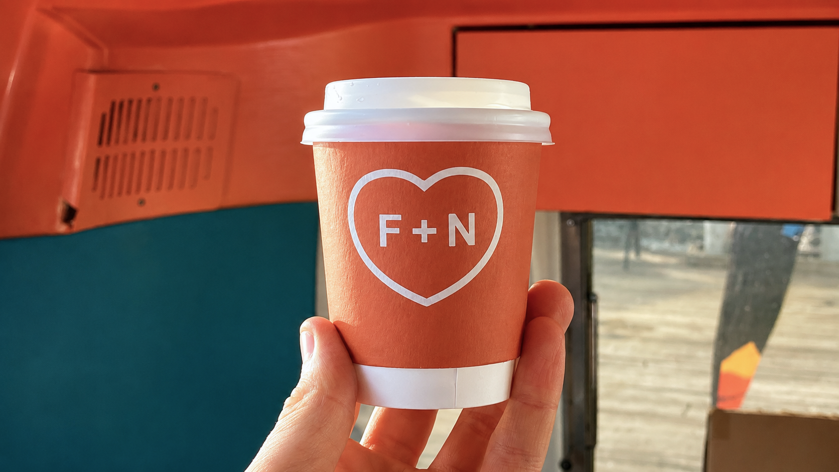 A coral takeaway cup with the F+N heart mark, held against the warm interior of Fred + Nancy’s.