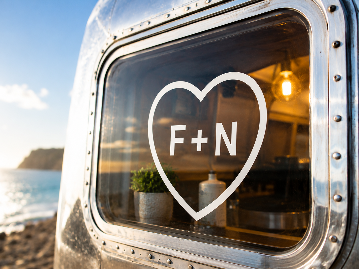 The F+N heart mark on the Airstream window at golden hour, sea visible behind, warm Airstream interior glowing through.