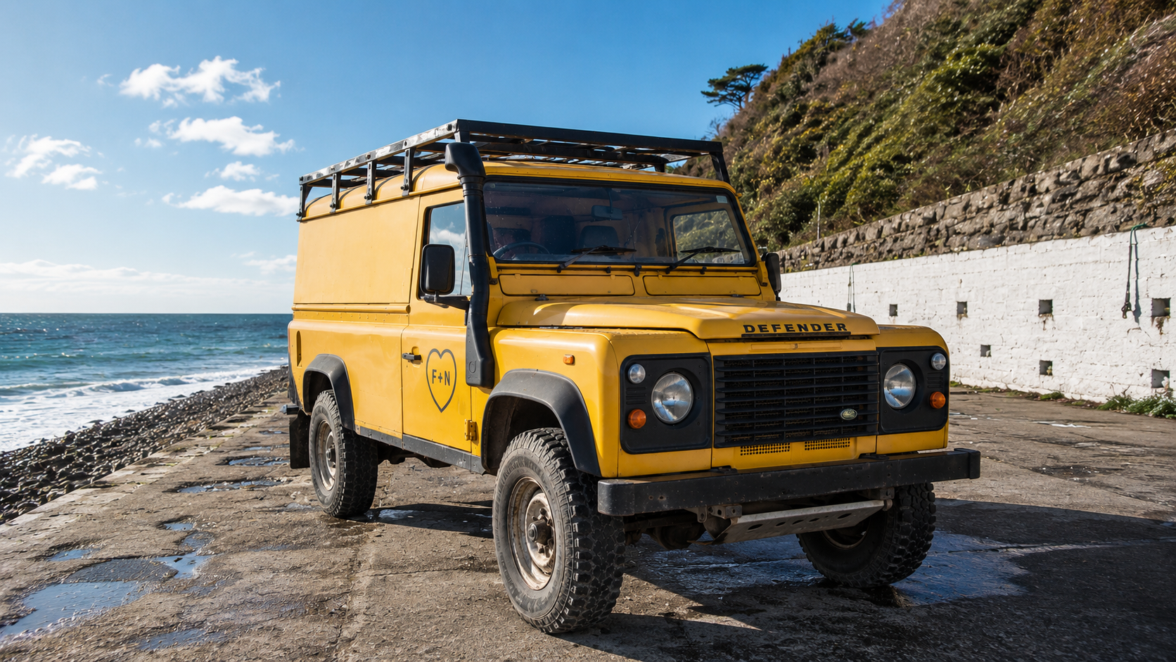 The Fred + Nancy's yellow Land Rover Defender parked on the seafront, F+N heart decal on the door, cliffs and sea behind.