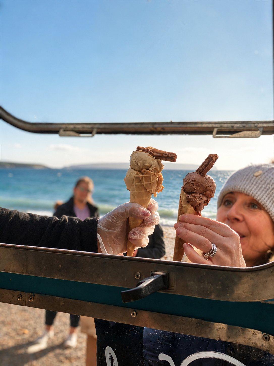 A gloved hand holds two ice cream cones with chocolate flakes, lifted through the Fred + Nancy's Airstream hatch at golden hour, sea behind.