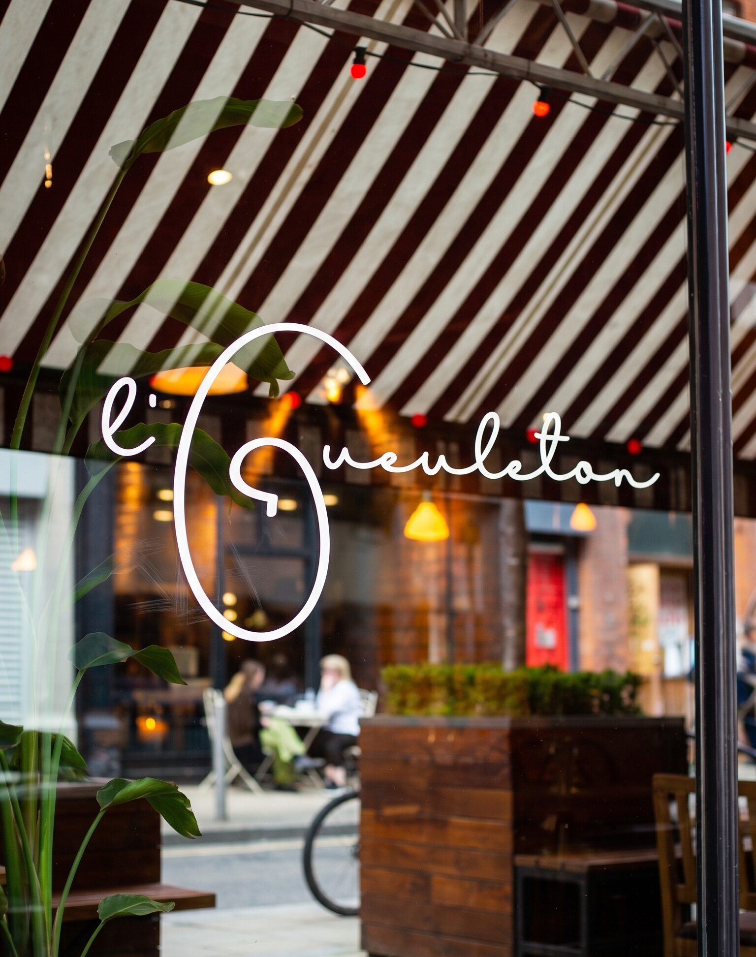 L'Gueuleton window decal with the white handwritten logo, striped awning behind.