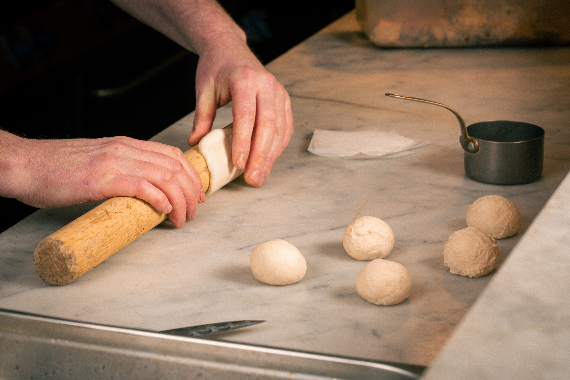 Hands rolling dough on a marble counter with small dough balls nearby.