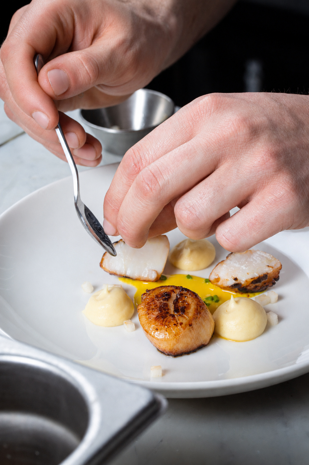 Chef's hands plating a scallop dish with sauce and garnish on a white plate.
