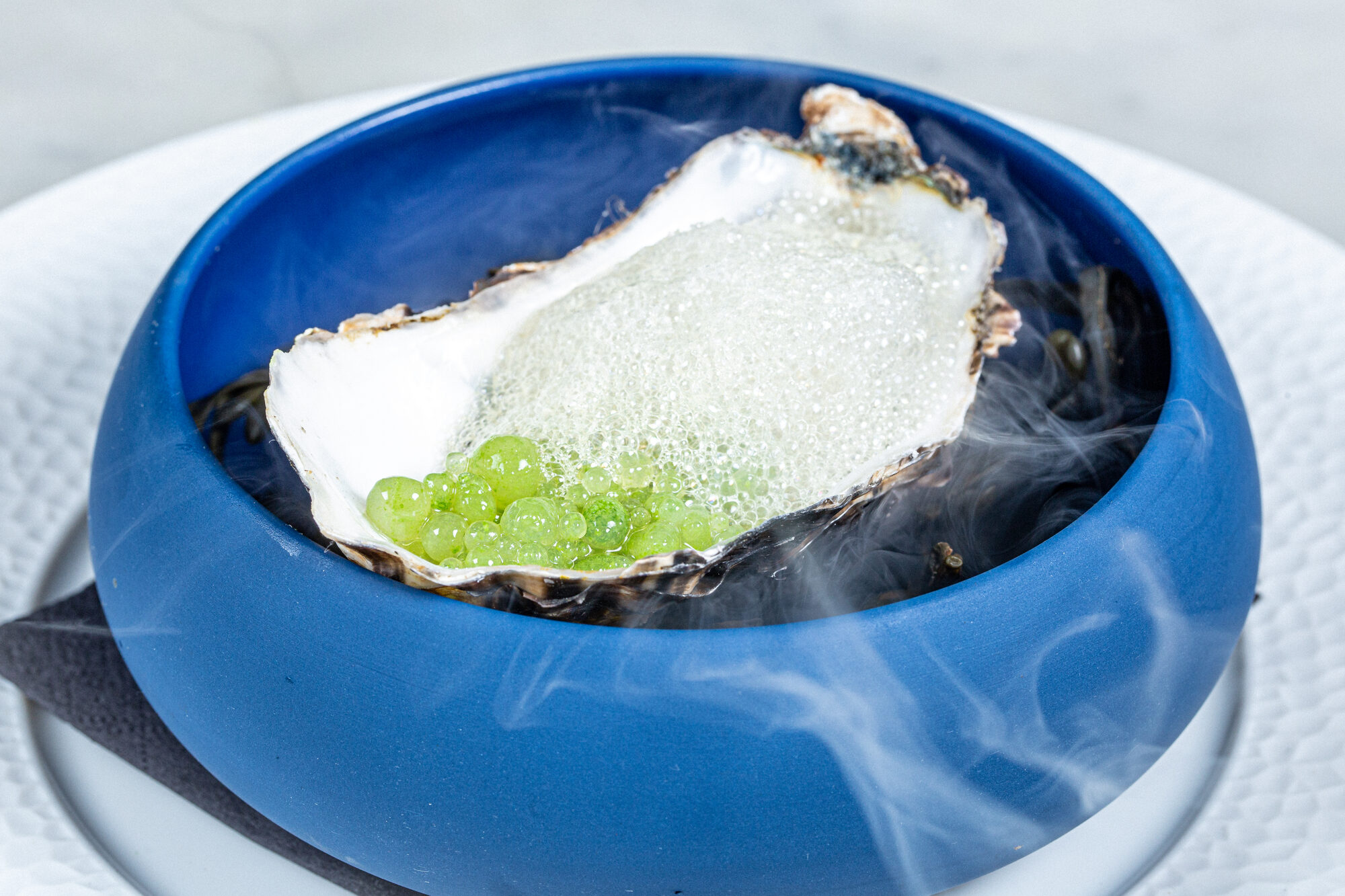 An oyster shell in a blue bowl with green garnish and visible smoke.