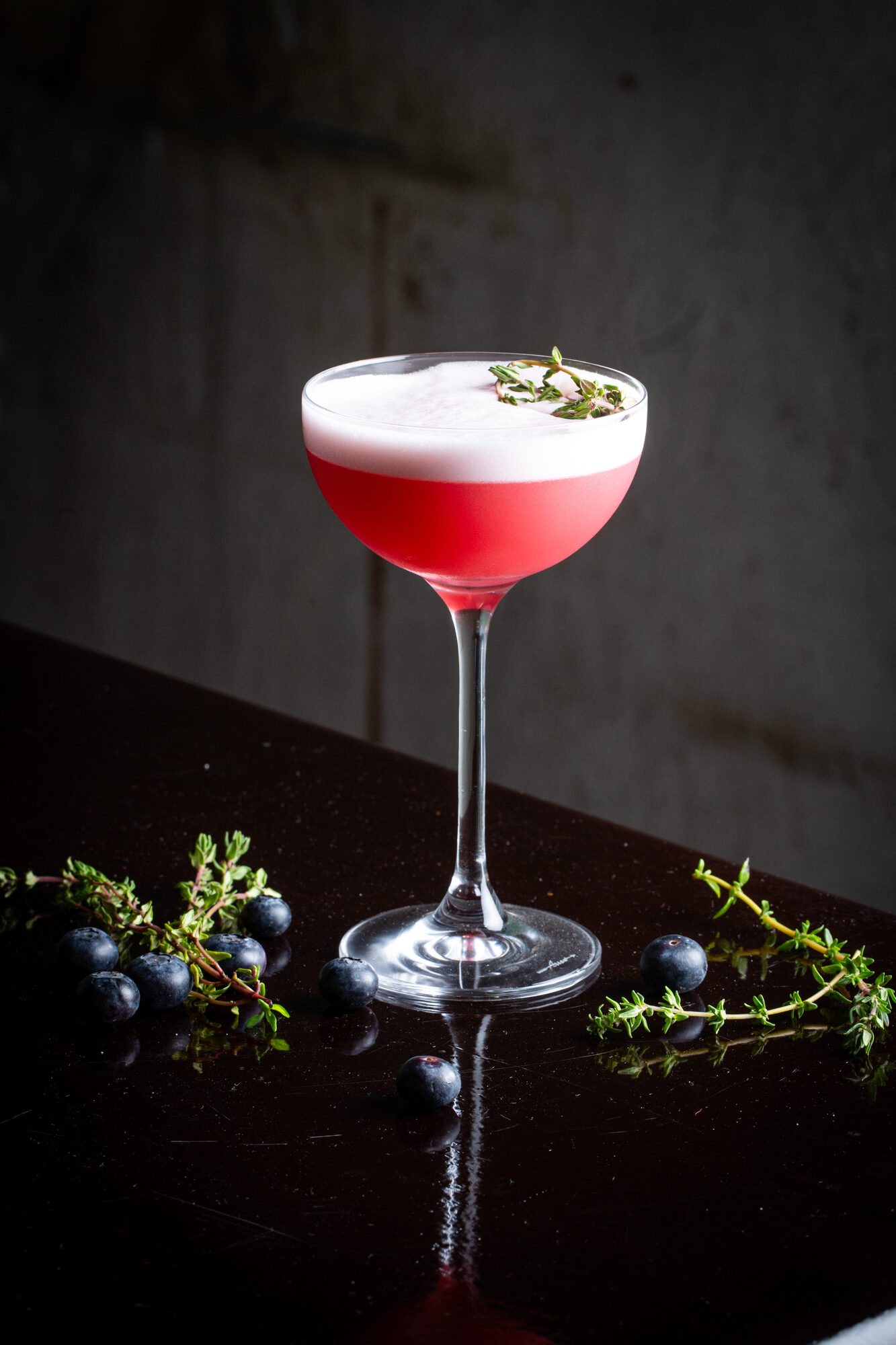 A red cocktail in a coupe glass with herbs and berries on a dark tabletop.