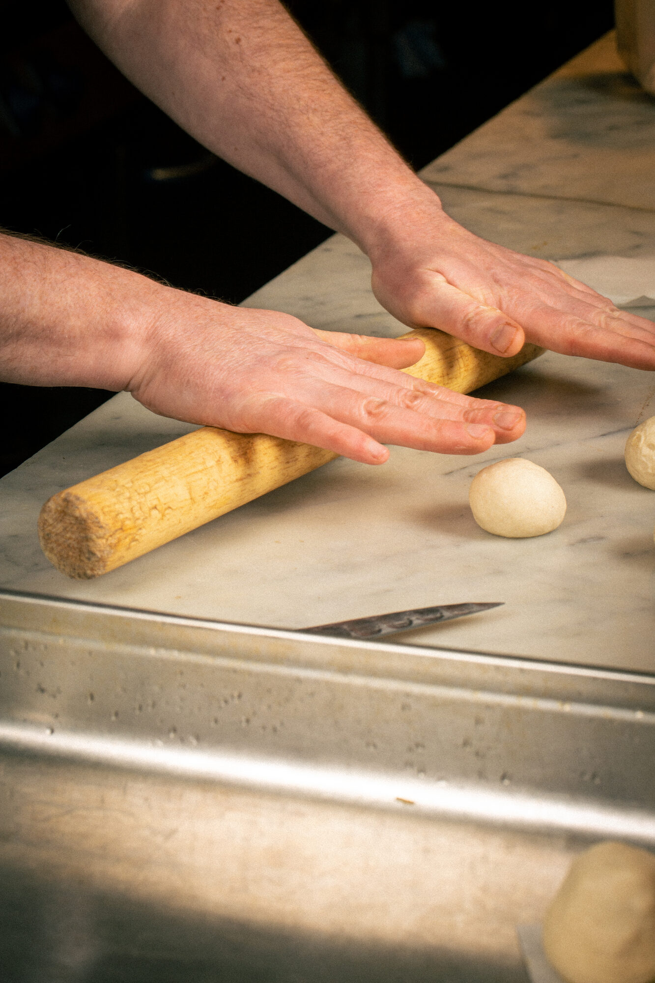 Hands rolling dough on a marble counter with small dough balls nearby.