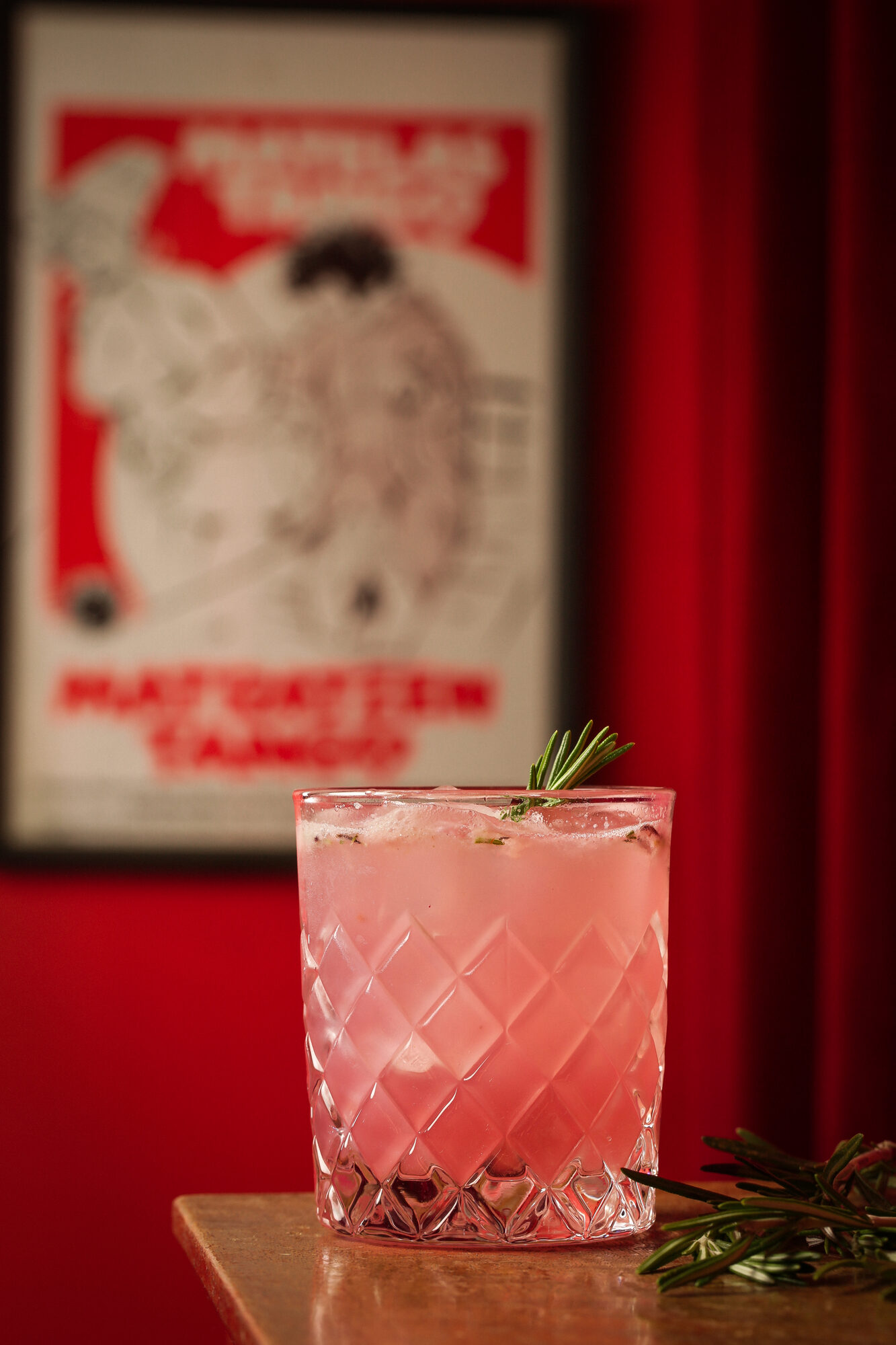 The Two Brothers cocktail — pink, rosemary-garnished, in a faceted rocks glass on a red velvet table, with a graphic poster on the red wall behind.