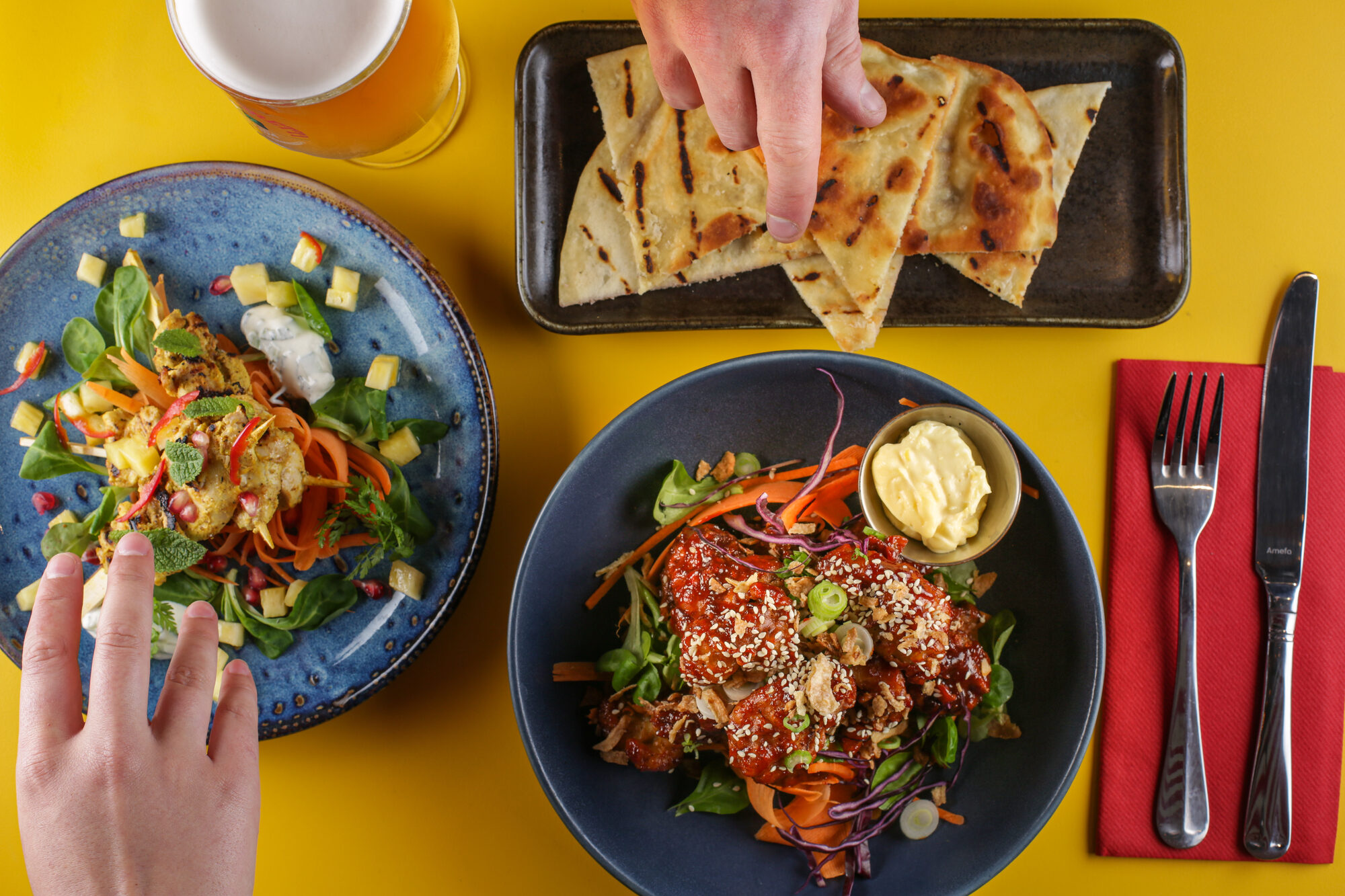 Overhead spread of food on a yellow table with hands reaching in.