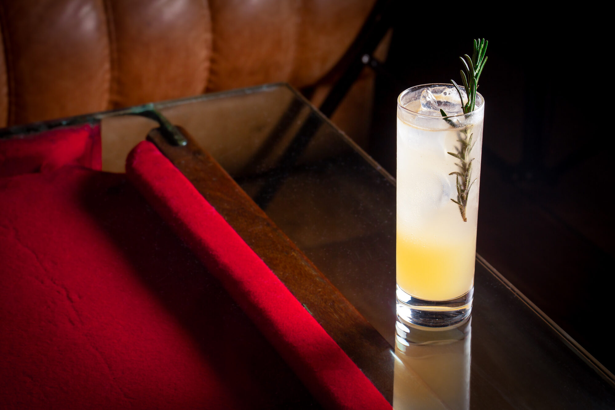 A highball cocktail on a glass tabletop with red velvet and tan leather behind at The Bar With No Name.